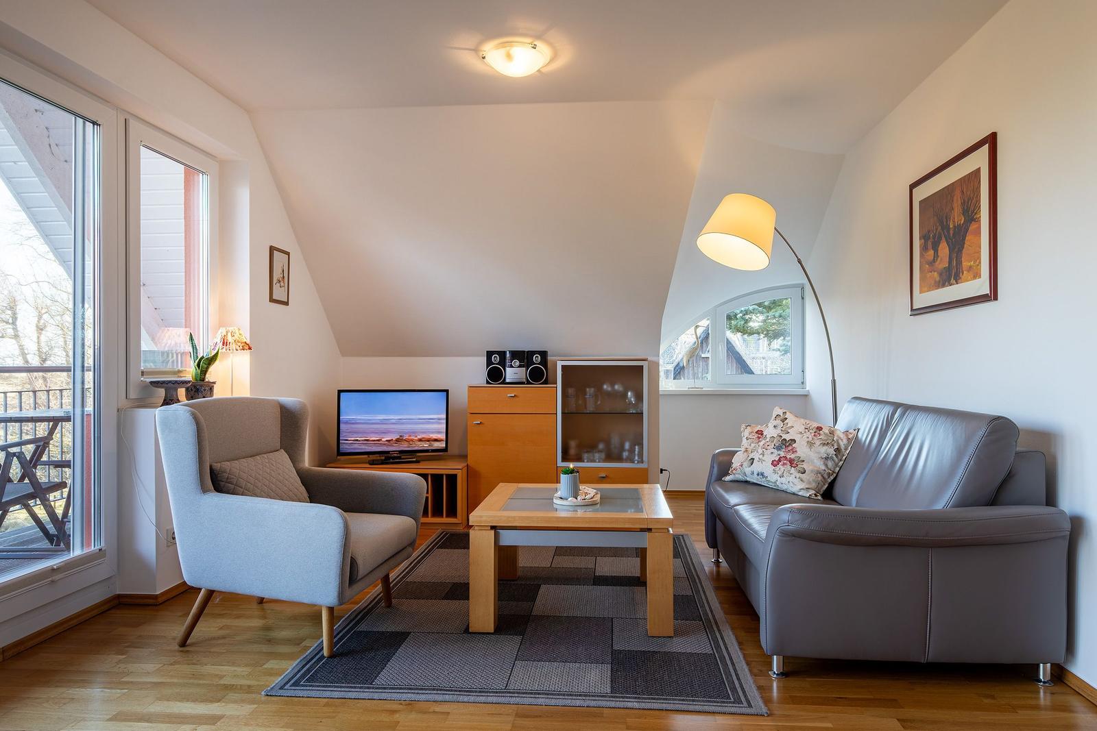 Wohnzimmer mit Sofa, Sessel, TV und Fenstern. Holzfußboden und Deckenlicht.