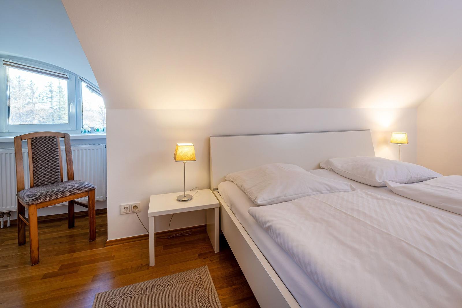 Schlafzimmer mit Doppelbett, Nachttisch und Stuhl. Fenster mit Blick auf Wald.