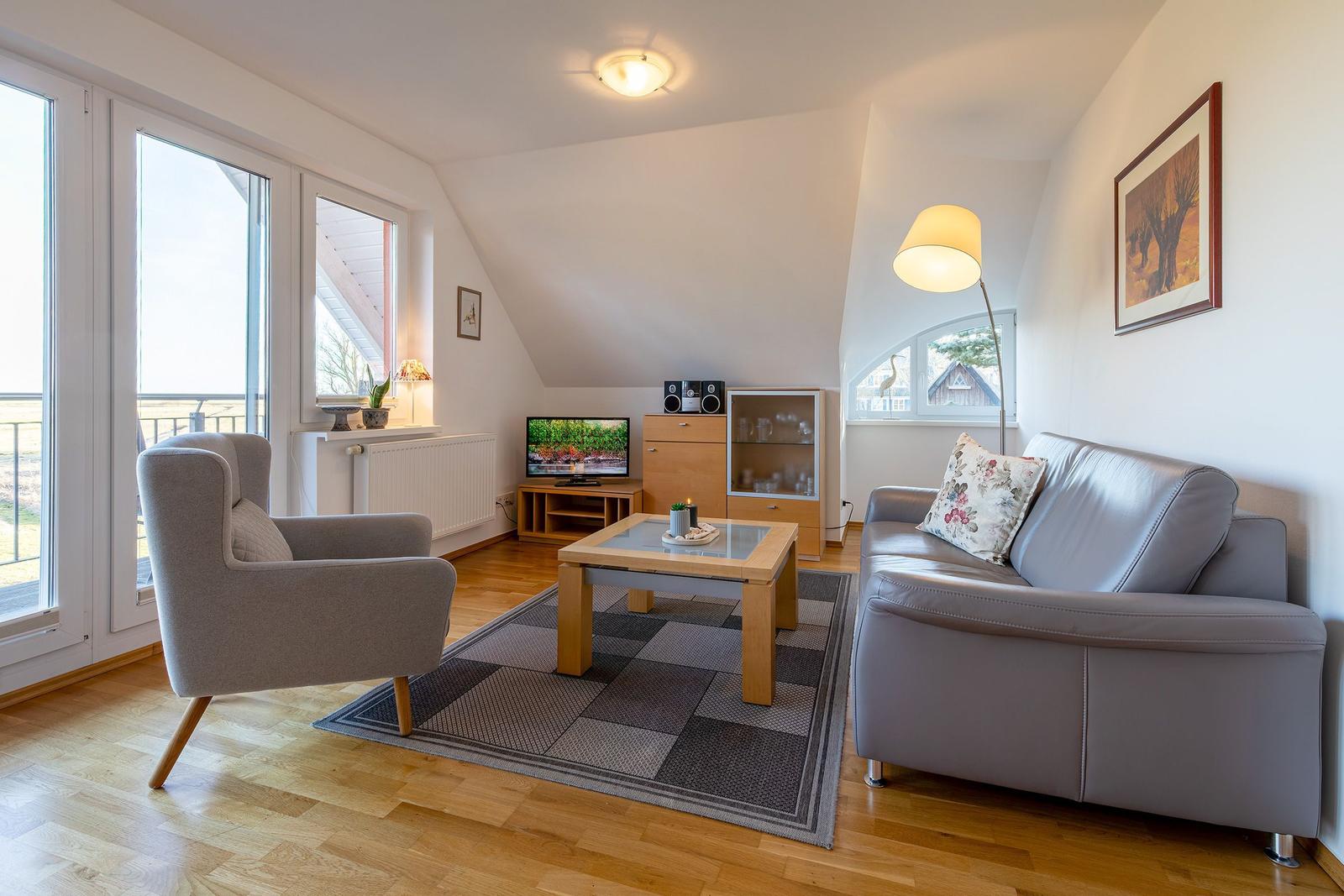 Wohnzimmer mit Sofa, Sessel, Couchtisch und TV. Holzboden und große Fenster.