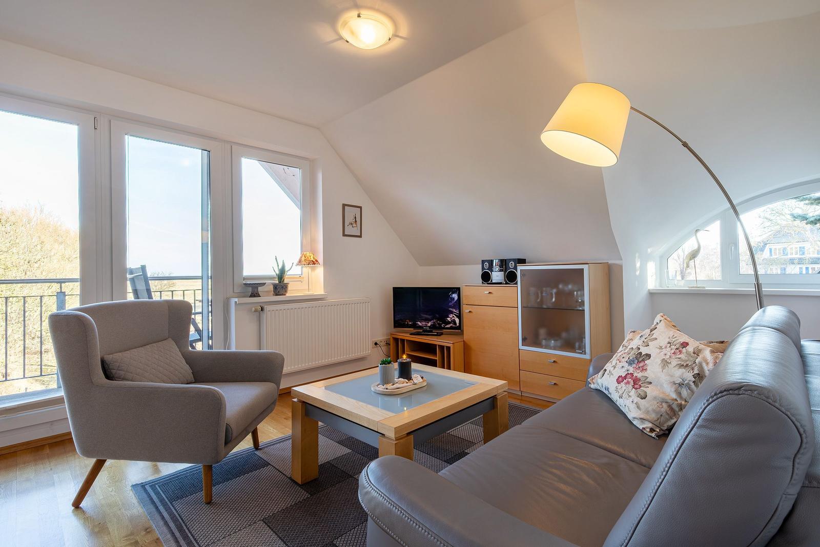 Wohnzimmer mit Sofa, Sessel, Couchtisch und TV. Große Fenster und Balkon. Holzboden und Schrank.