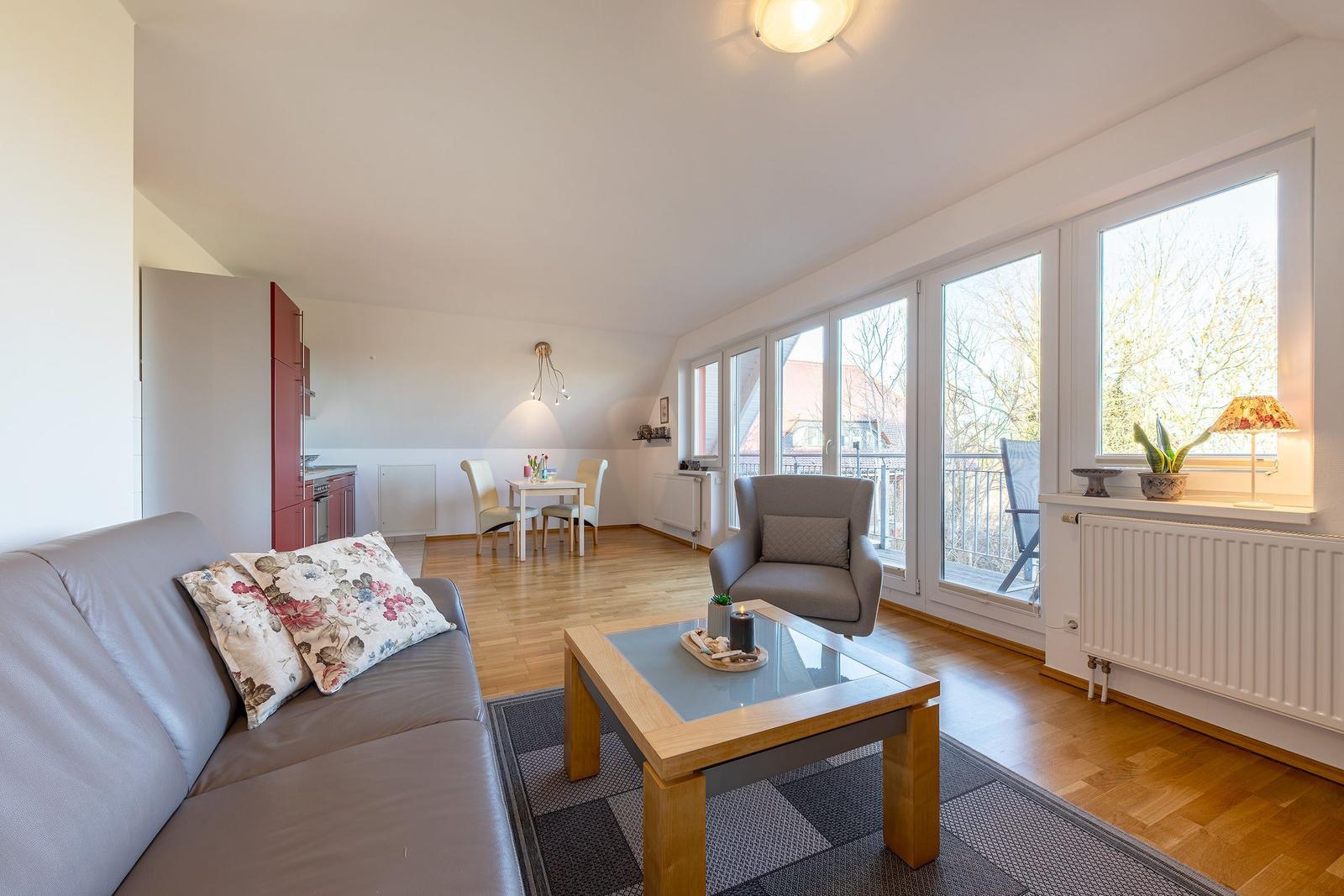 Wohnbereich mit Sofa, Sessel und Esstisch. Durchgang zur Küche. Große Fensterfront mit Blick auf Balkon.