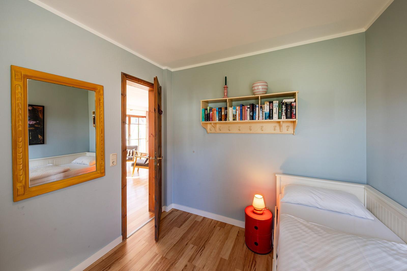 Schlafzimmer mit Bett, Wandregal und Spiegel. Tür führt in Wohnbereich.