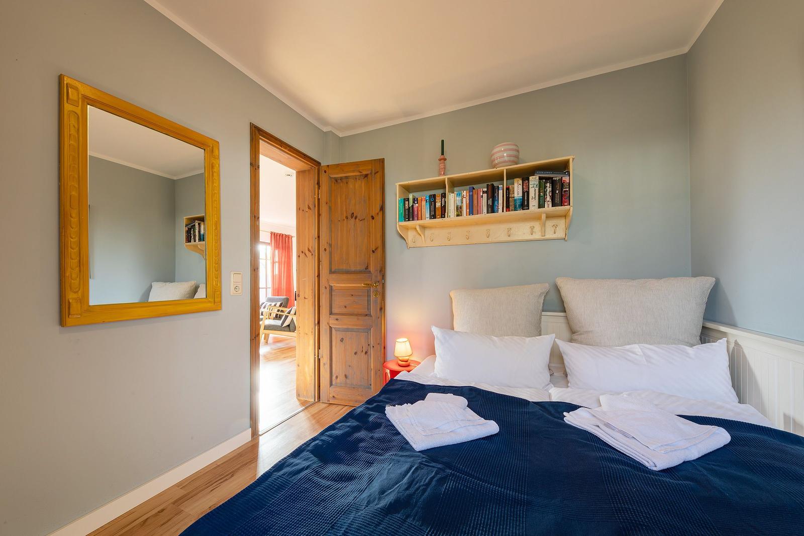 Schlafzimmer mit Bett, Spiegel und Bücherregal. Tür führt in angrenzenden Raum.