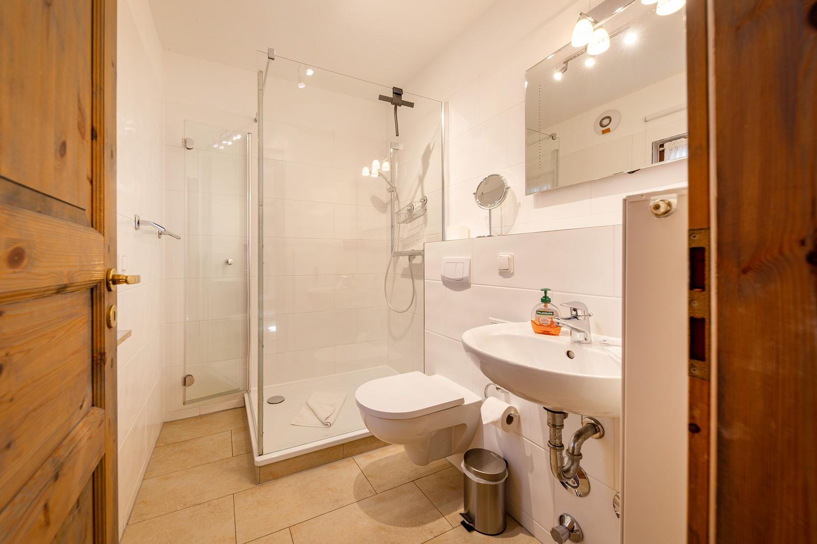 Bad mit Dusche, Waschbecken und Toilette. Holztür und weiße Fliesen.