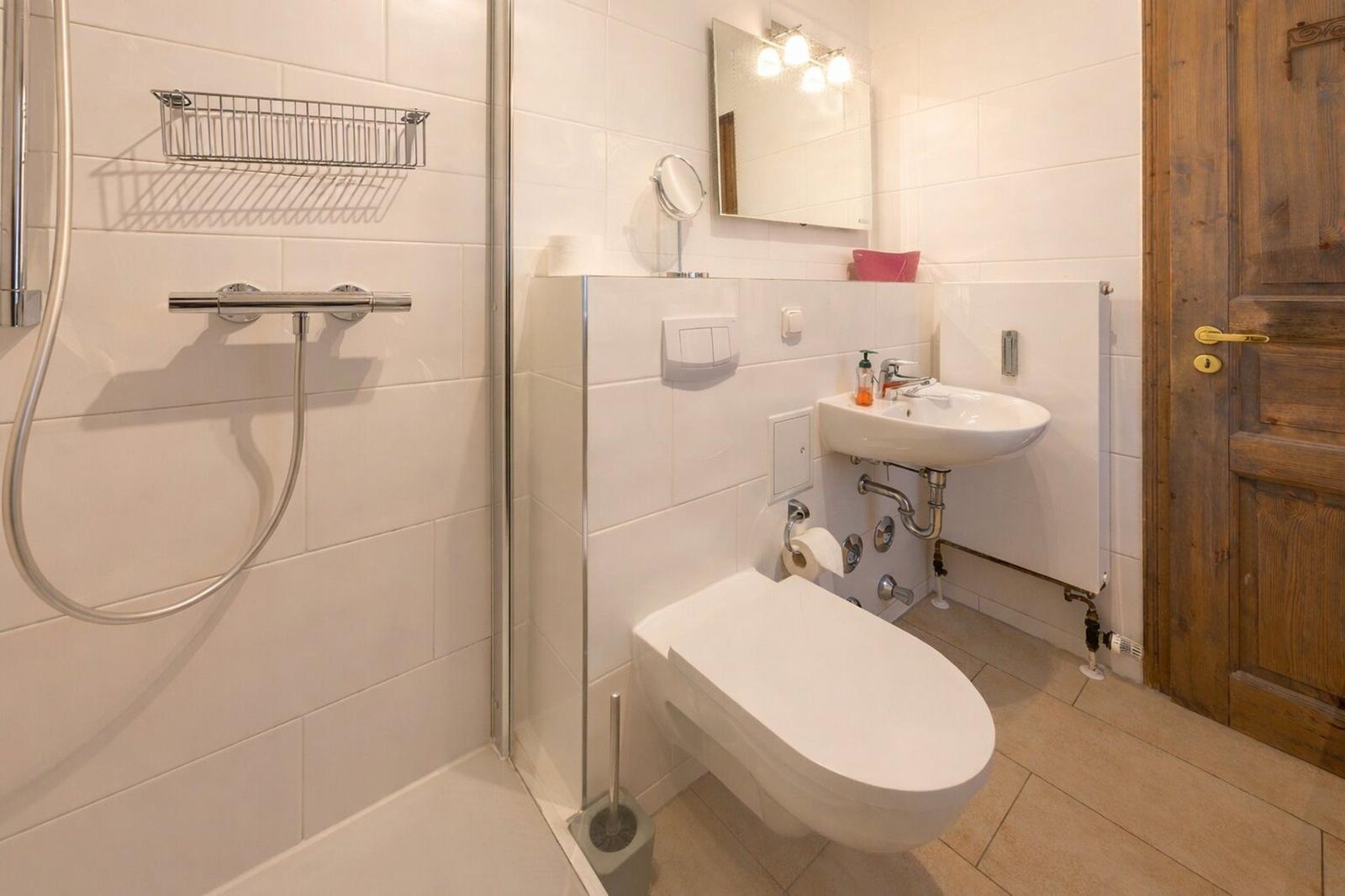 Badezimmer mit Dusche, Waschbecken und Toilette. Holztür und weiße Fliesen.