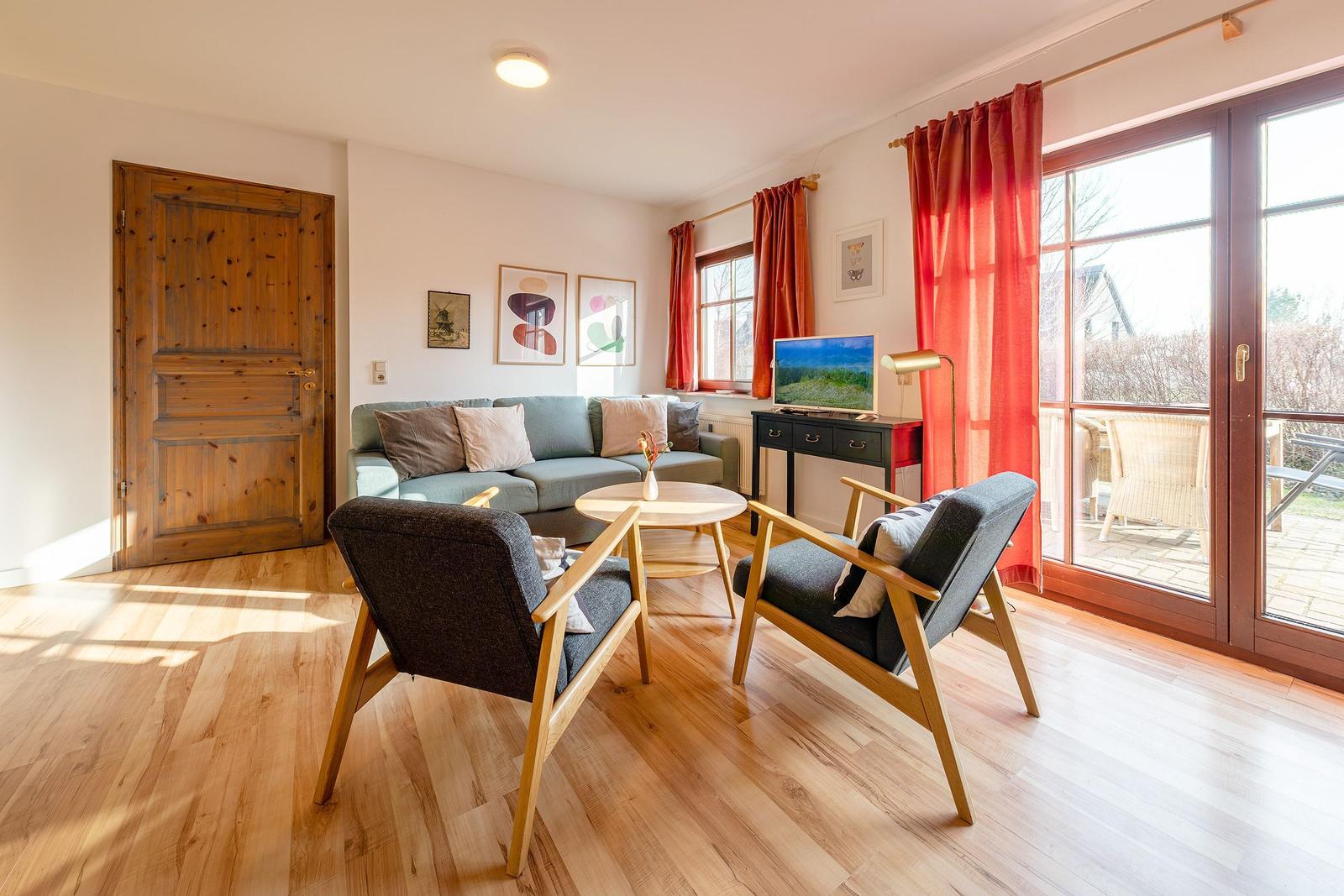 Wohnzimmer mit Sofa, zwei Stühlen und TV. Holzboden und große Fenster.