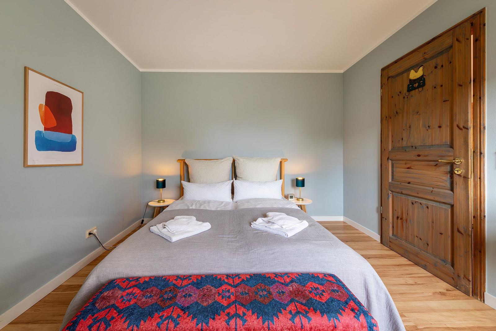 Schlafzimmer mit Bett, zwei Nachttischen und Holztür. Wandbild und gemusterte Decke.