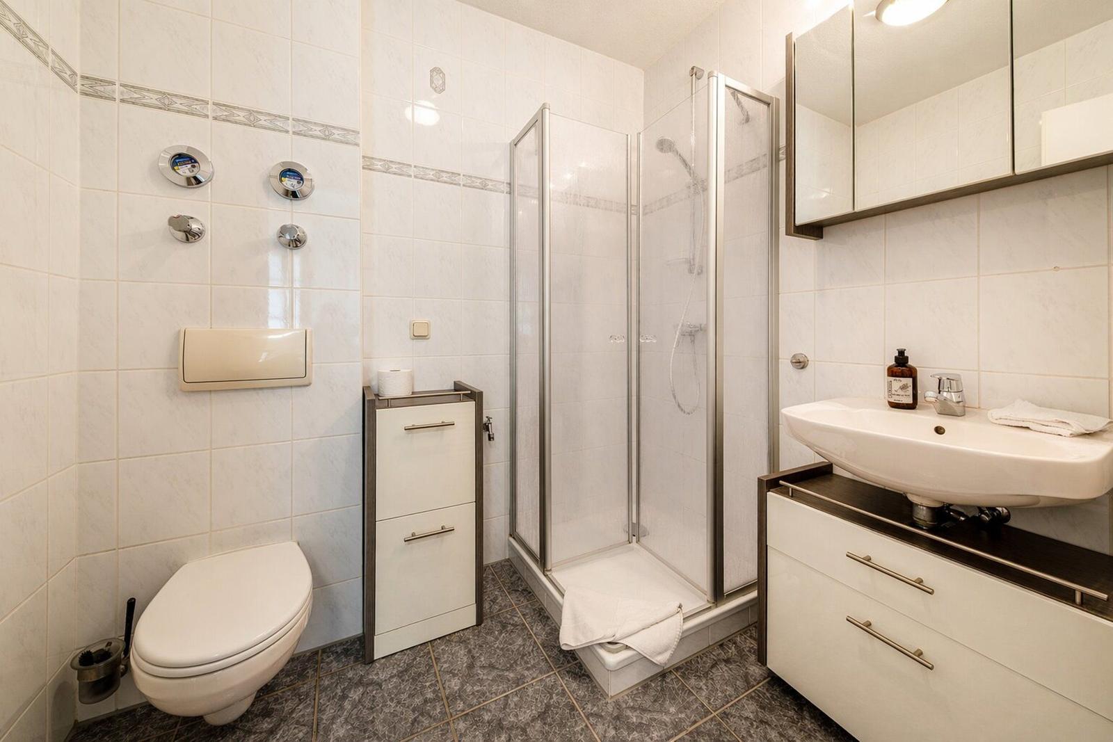 Badezimmer mit Dusche, Waschbecken und Toilette. Weiße Fliesen und dunkler Boden.