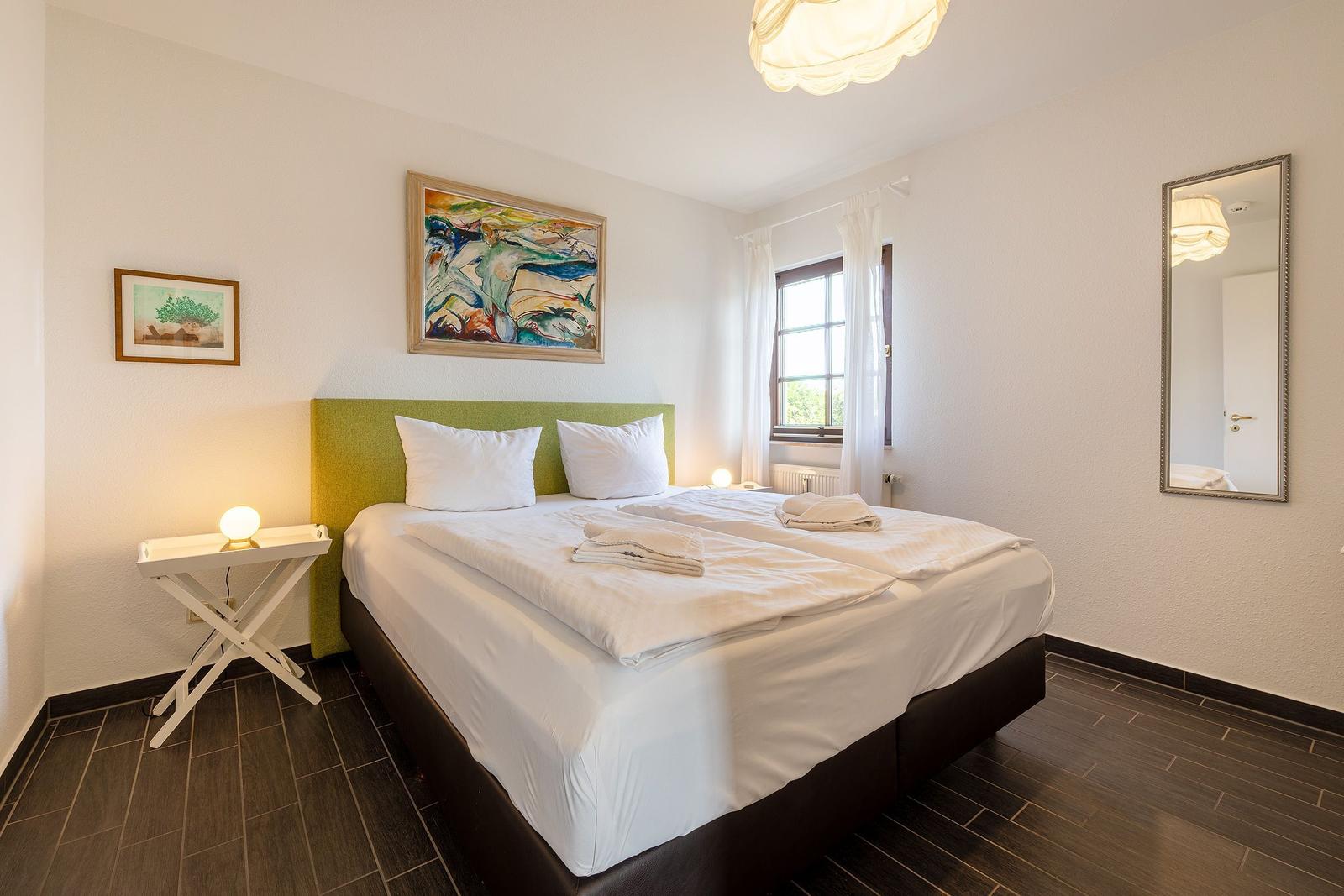 Schlafzimmer mit Doppelbett, grünem Kopfteil, Fenster und Wandspiegel.