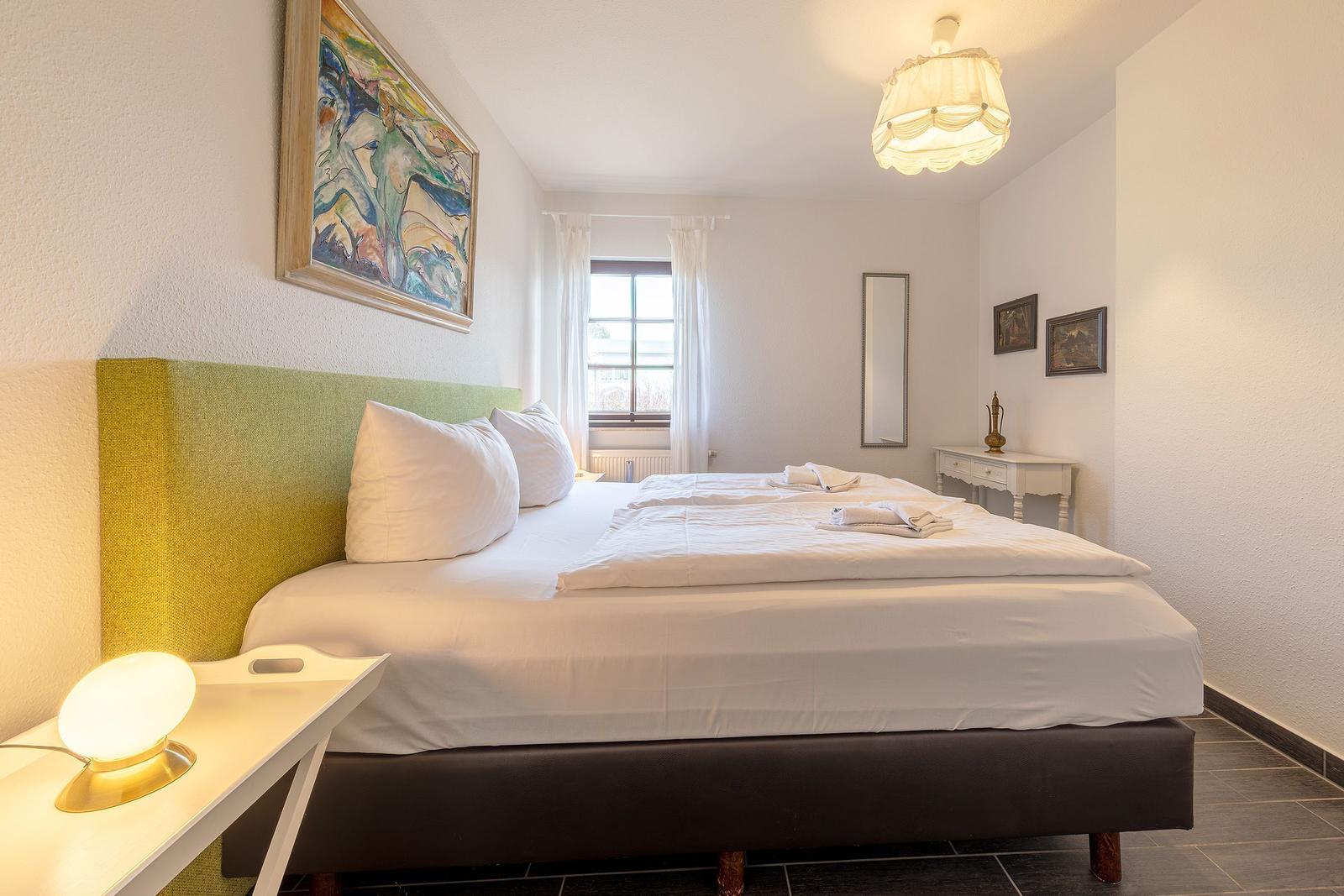 Schlafzimmer mit Bett, Fenster, Wandbild und Nachttisch mit Lampe.