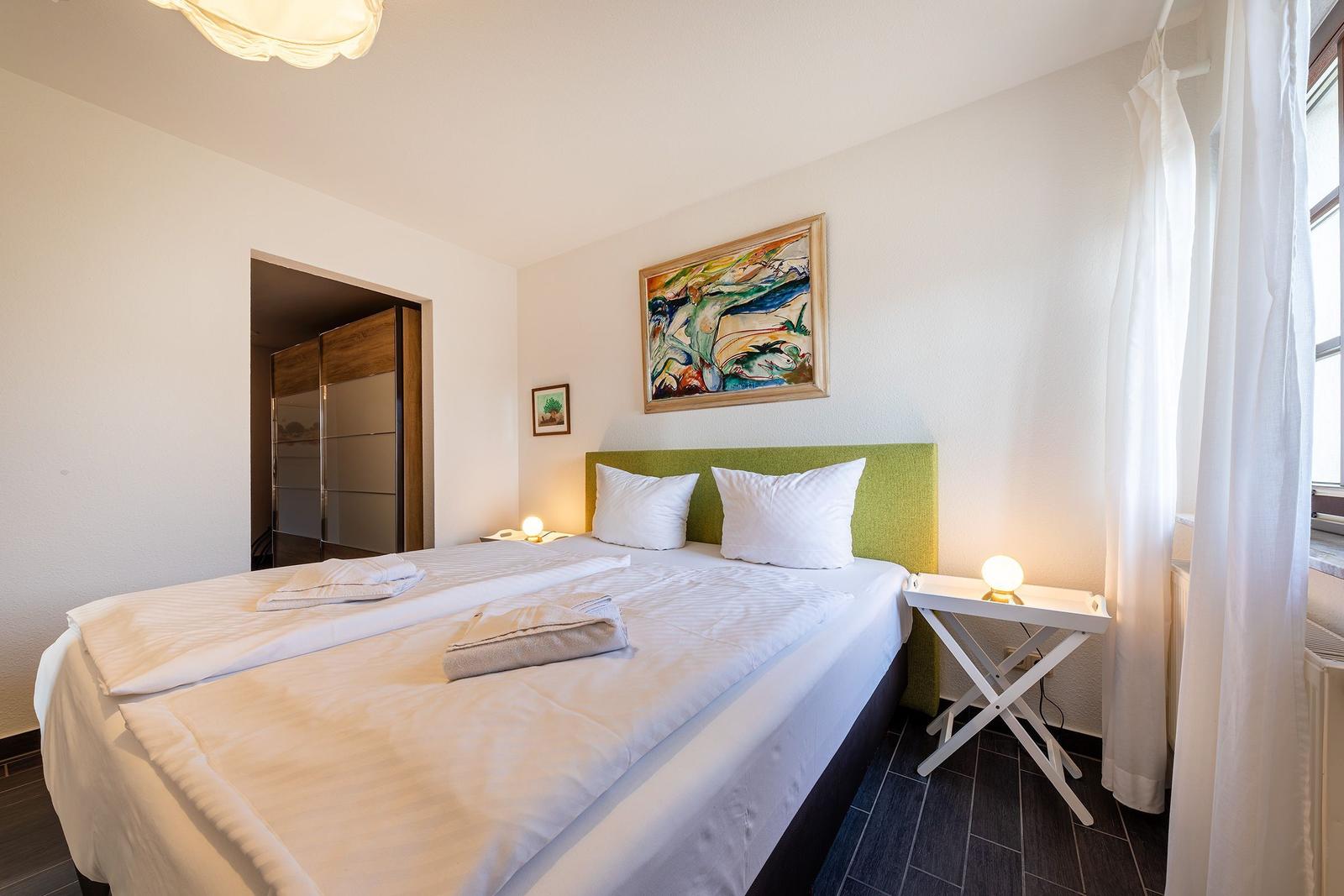 Schlafzimmer mit Doppelbett, grünem Kopfteil und Wandbild. Neben dem Bett steht ein kleiner Tisch.