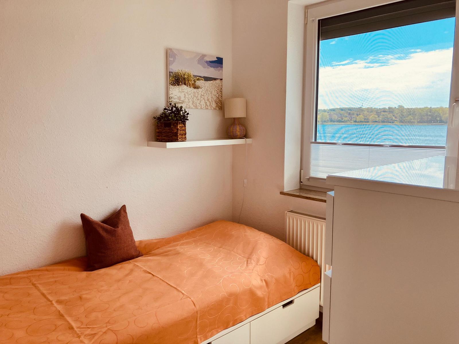 Schlafzimmer mit Blick auf See, Bett mit orangefarbener Bettwäsche und Fenster mit Jalousie.