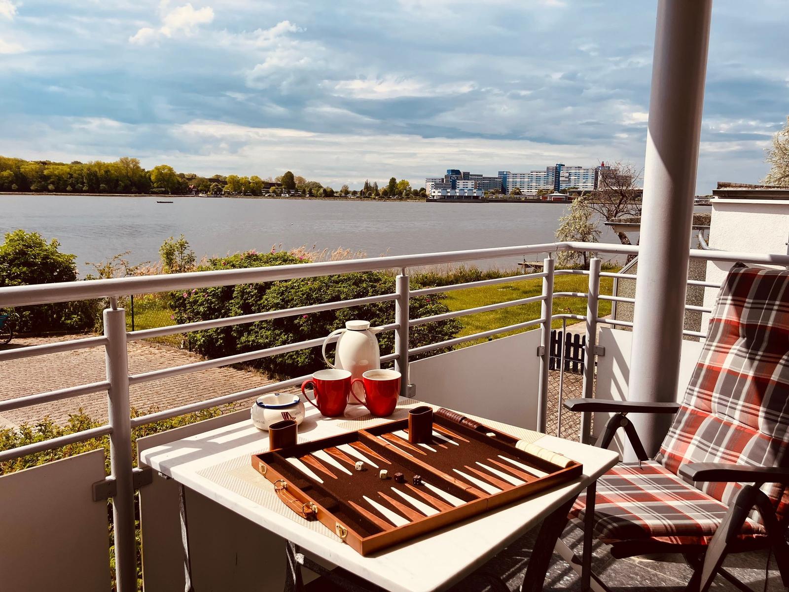 Balcony with table, backgammon, cups, and chair overlooking river and cityscape.