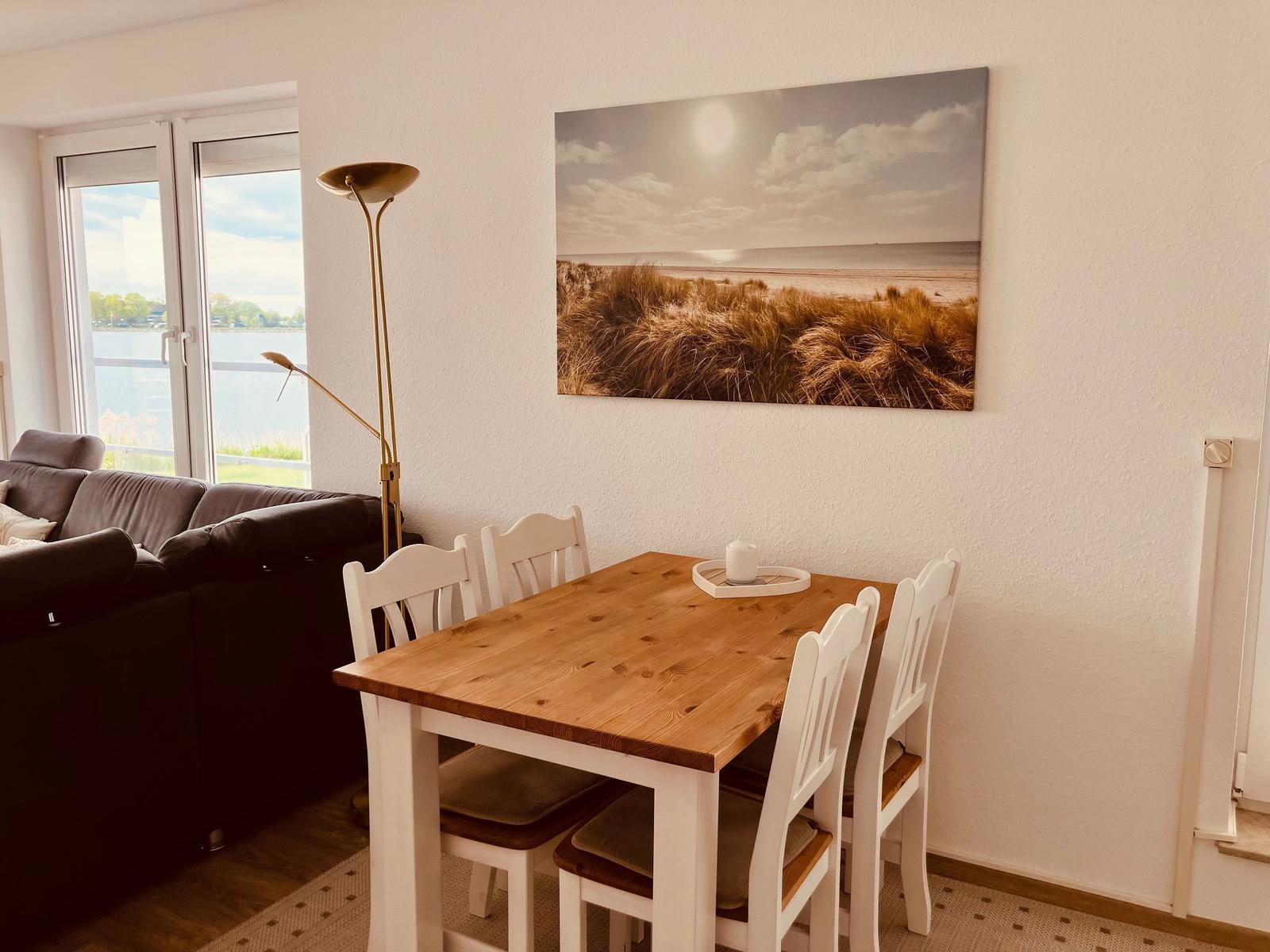 Essbereich mit Holztisch und weißen Stühlen. Wandbild zeigt Strandlandschaft. Fenster mit Blick auf Wasser.