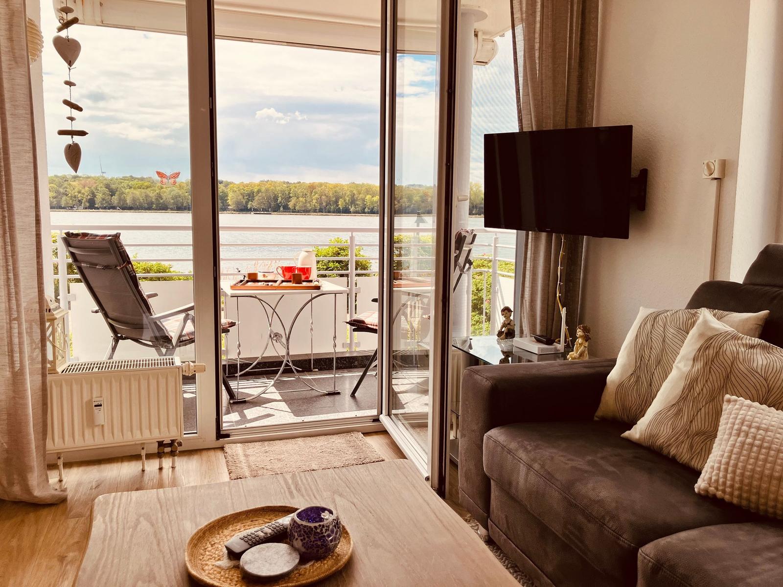 Wohnzimmer mit Balkon, Blick auf Fluss und Wald, Sofa, Fernseher und Tisch.