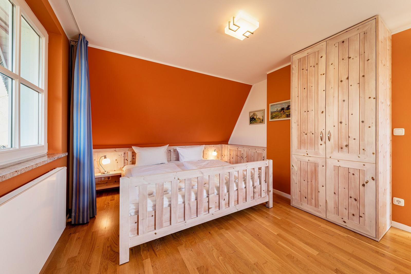 Schlafzimmer mit Holzbett, Kleiderschrank und Fenster. Wände orange, Holzboden.
