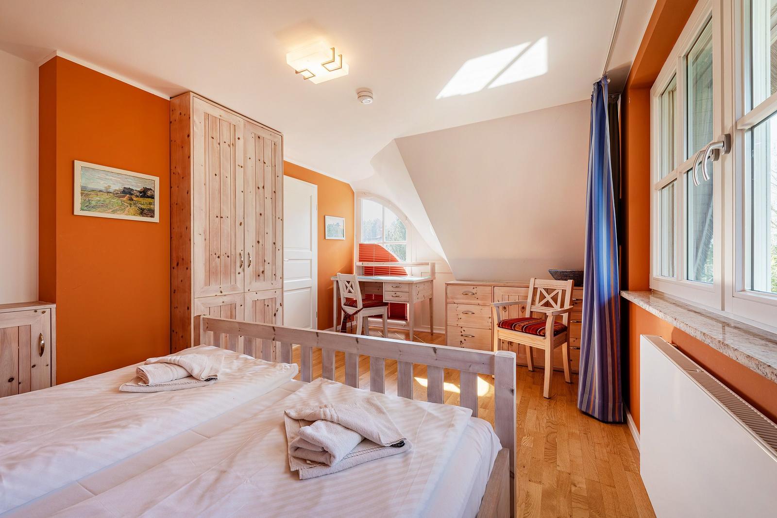 Schlafzimmer mit Doppelbett, Schreibtisch und Fenster. Holzmöbel und orange Wandfarbe.