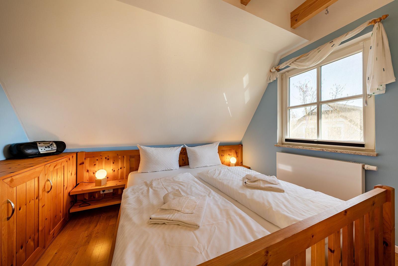 Schlafzimmer mit Doppelbett, Holzmöbel und Fenster. Decke geneigt, Licht durch Fenster.