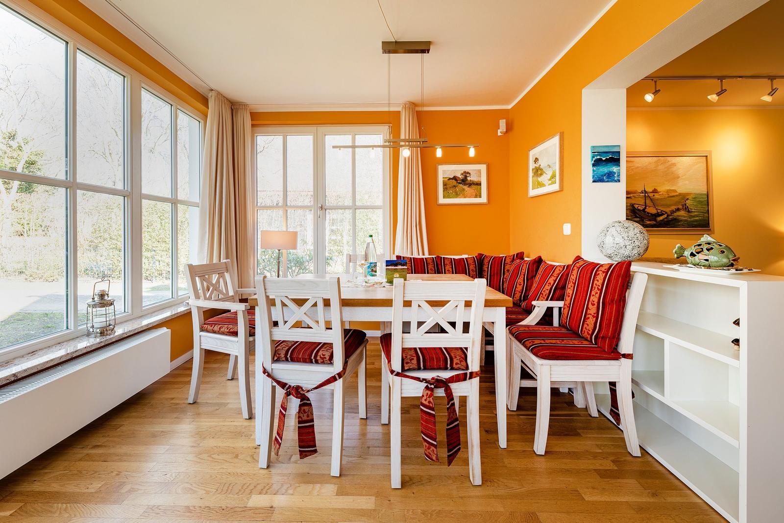 Essbereich mit weißem Tisch, Stühlen und Sitzbank mit roten Kissen. Große Fenster, Holzboden und orange Wände.
