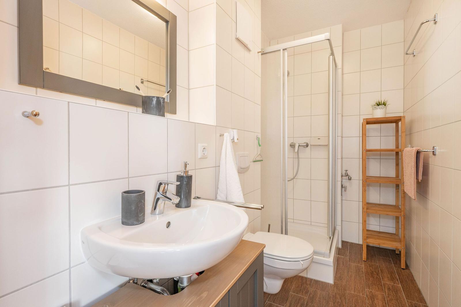 Badezimmer mit Dusche, Waschbecken und Toilette. Holzregal und Spiegel.