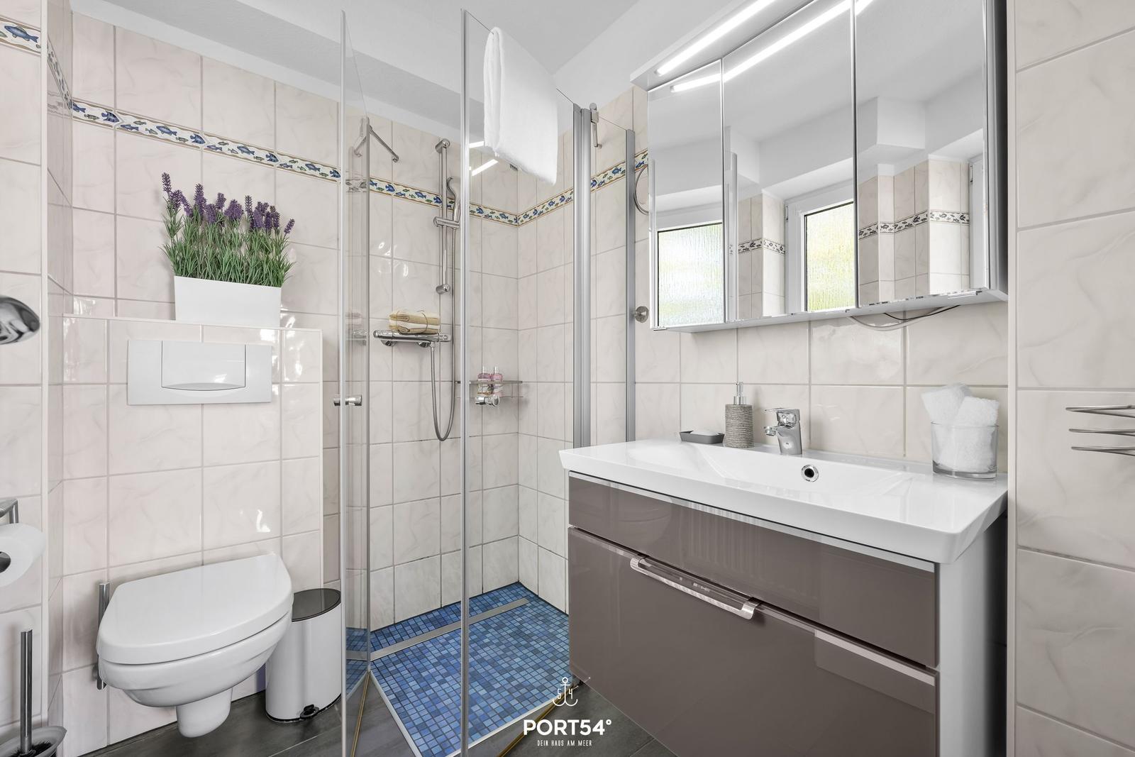 Badezimmer mit Dusche, Waschbecken und Toilette. Fliesen und Spiegel sind sichtbar.