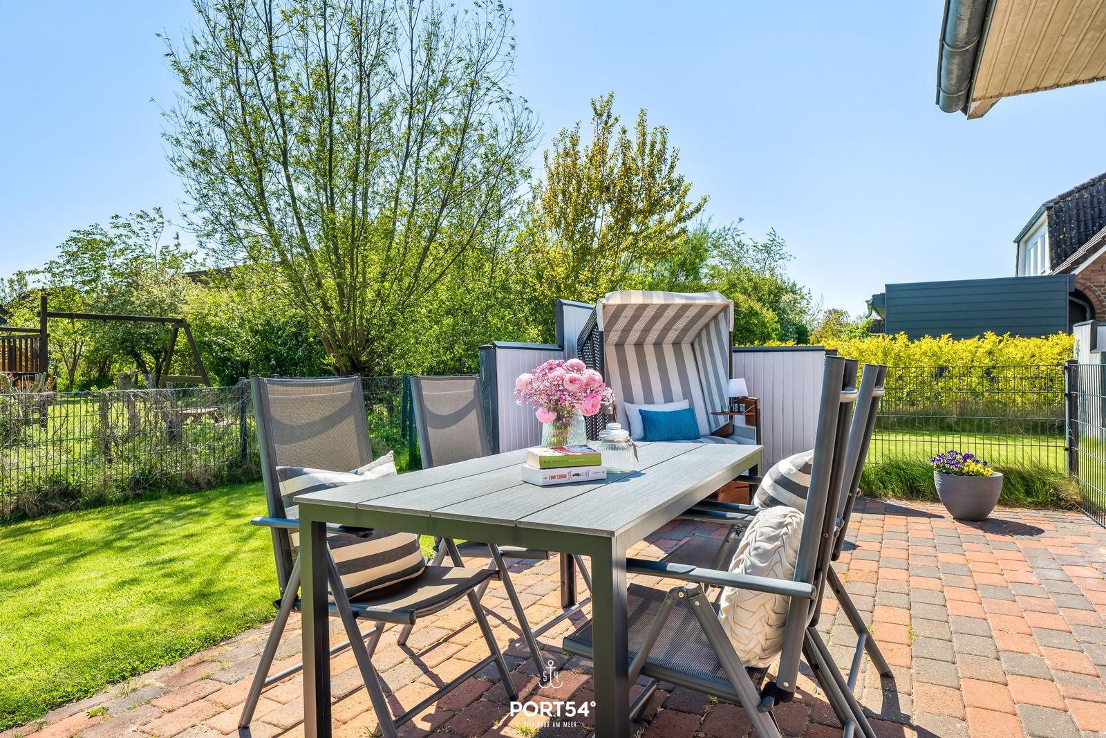 Terrasse mit Tisch, Stühlen und Strandkorb. Grünfläche und Zaun im Hintergrund.
