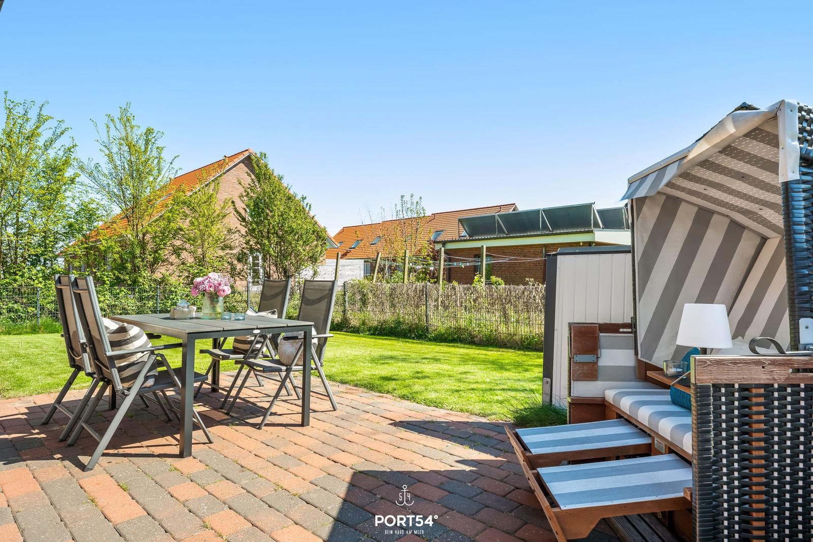 Terrasse mit Tisch, Stühlen und Strandkorb. Grünfläche und Häuser im Hintergrund.