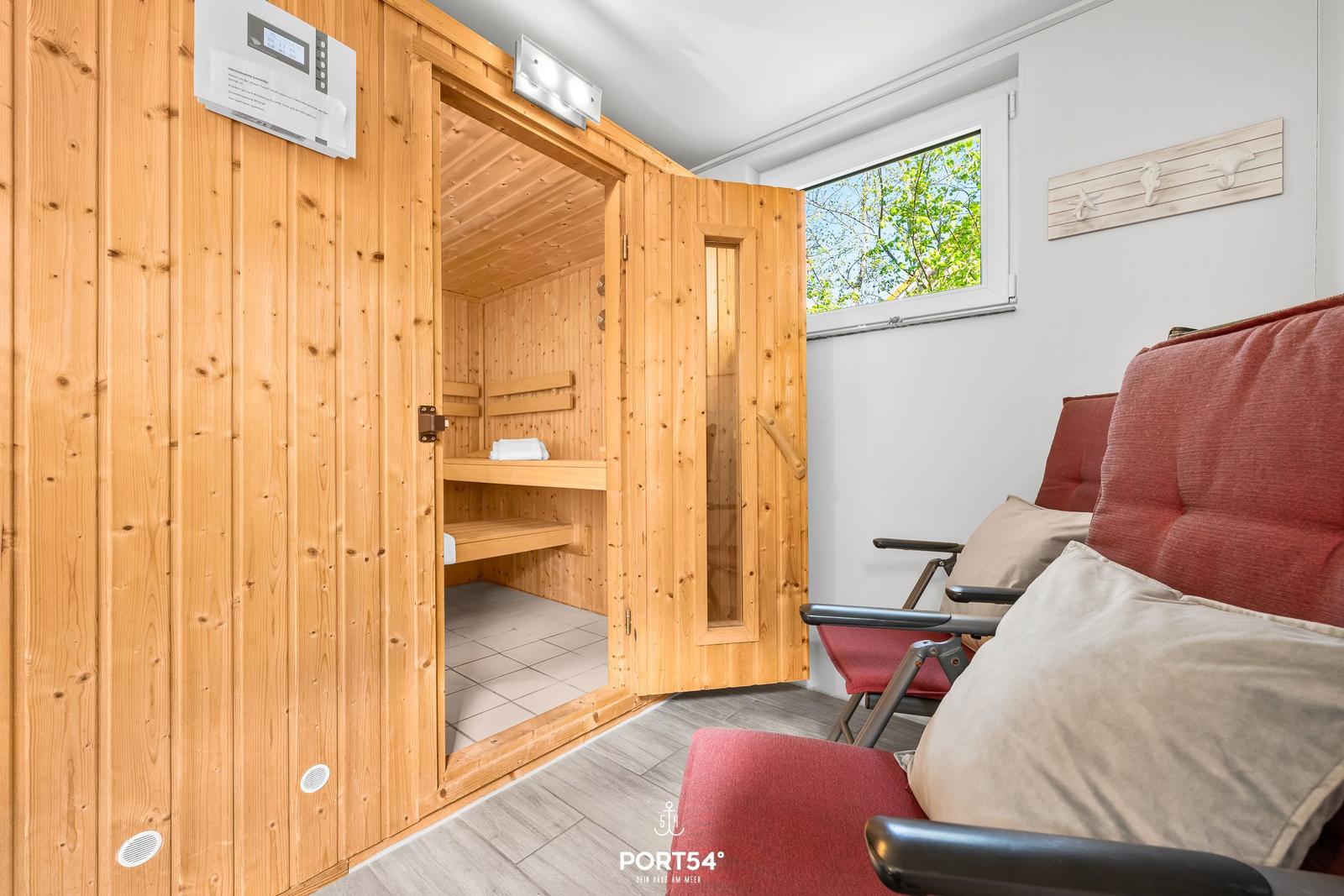 Sauna mit Holzverkleidung und Sitzbänken. Rote Sessel und Fenster mit Blick auf Bäume.