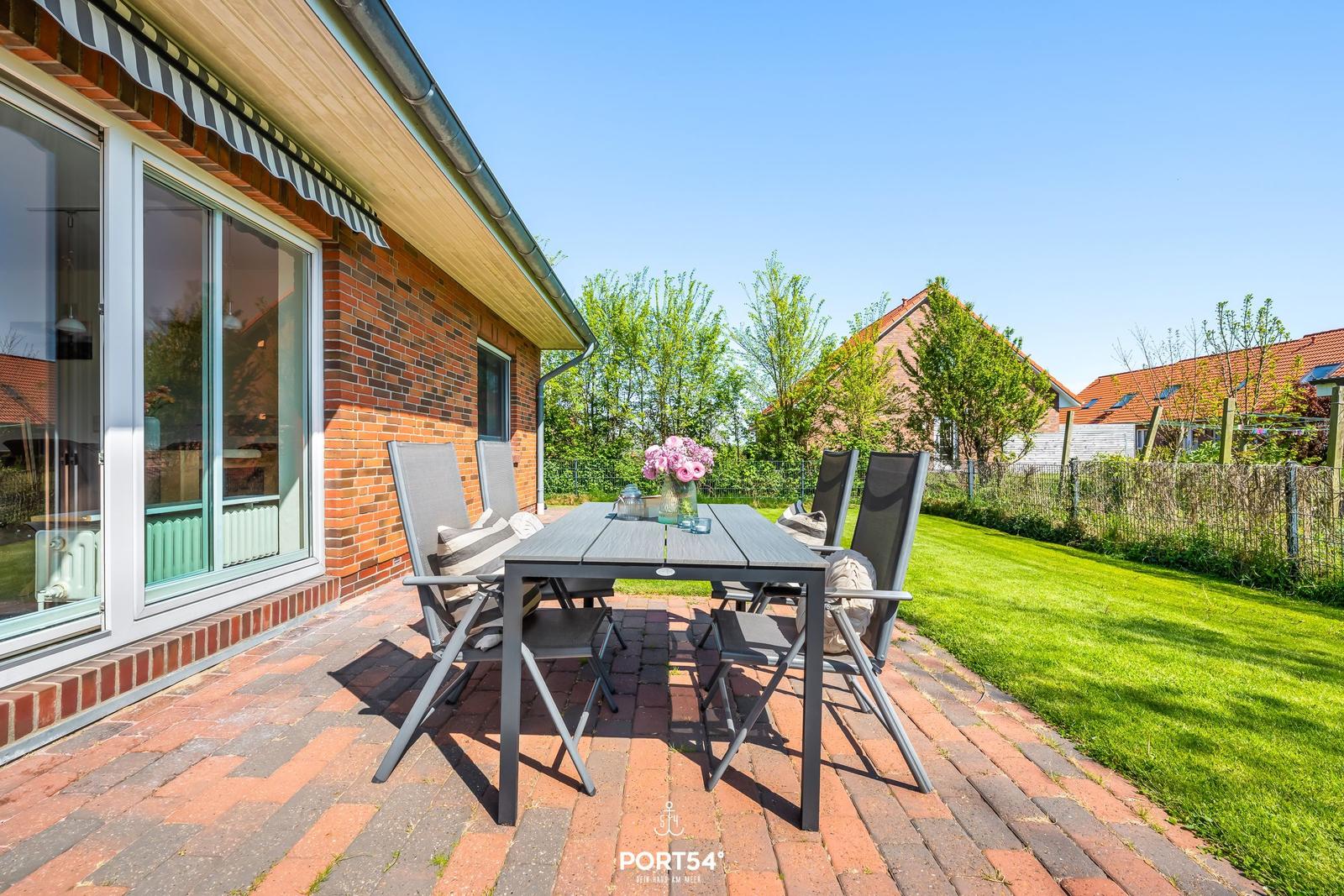 Terrasse mit Tisch und Stühlen, roter Pflastersteinboden, grüner Rasen und Blick auf Nachbarhäuser.