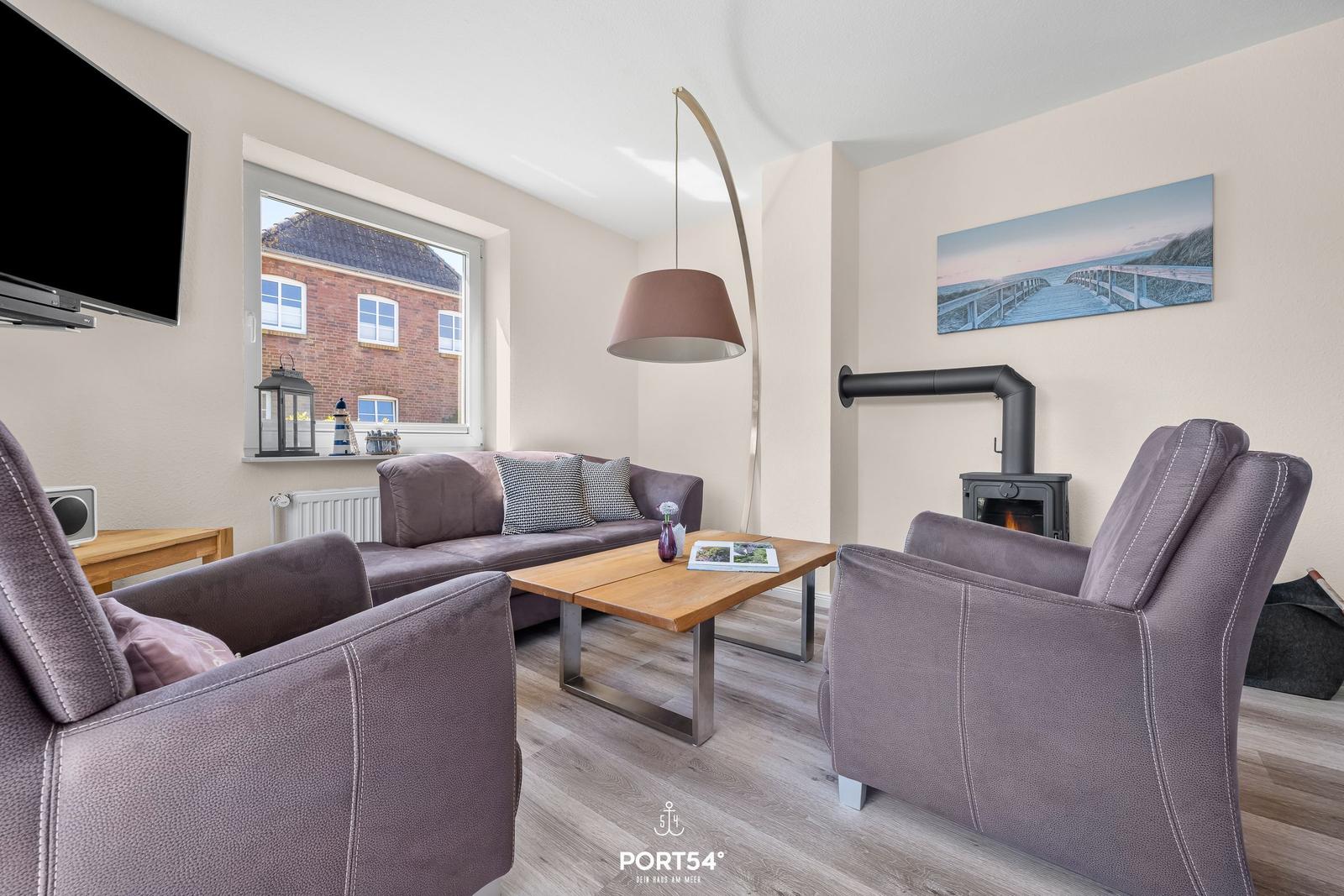 Wohnzimmer mit Sofa, zwei Sesseln, Kamin und Fernseher. Fenster zeigt Haus außen.