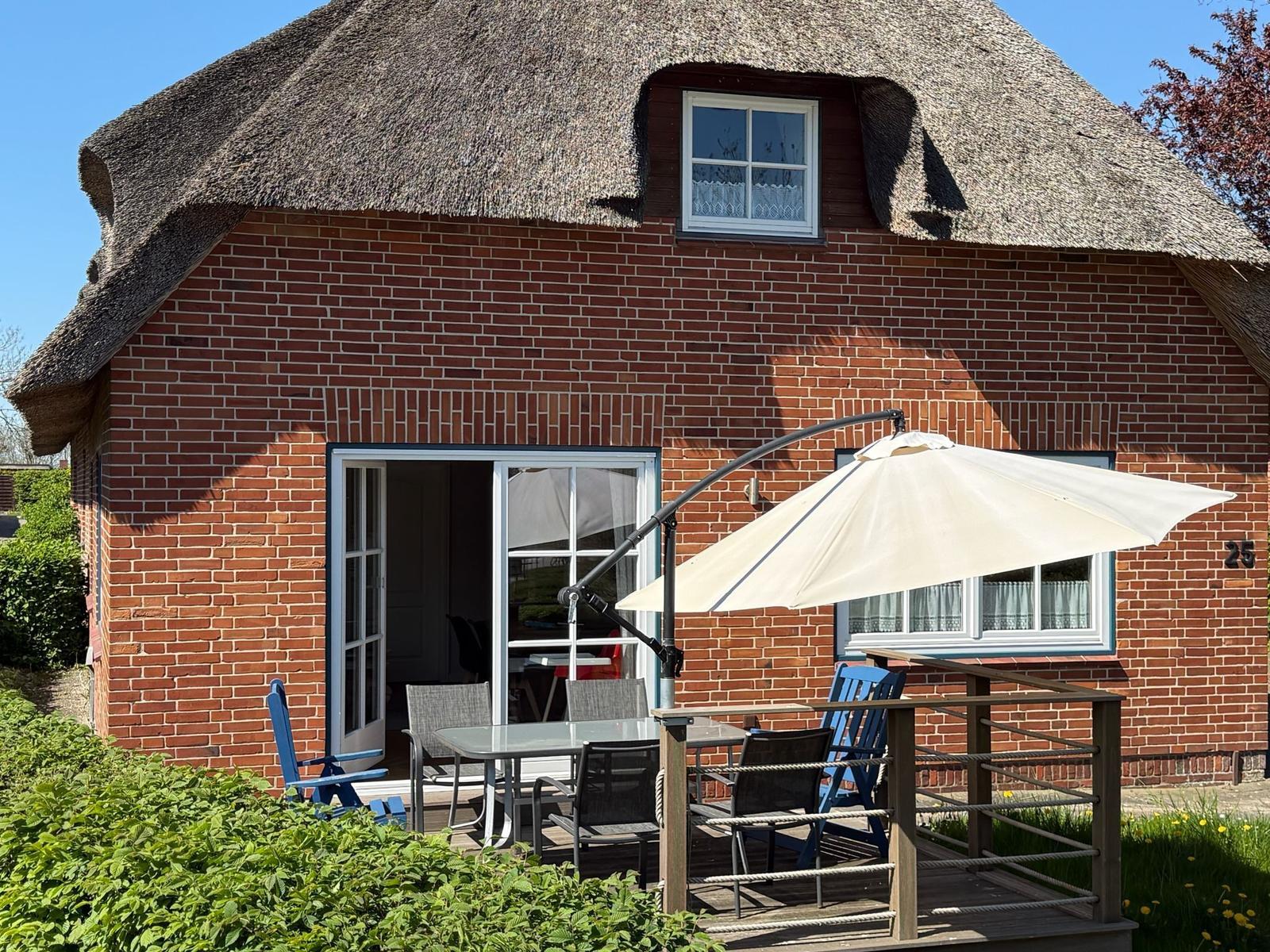 Rotes Backsteinhaus mit Strohdach, Terrasse mit Tisch, Stühlen und Sonnenschirm.