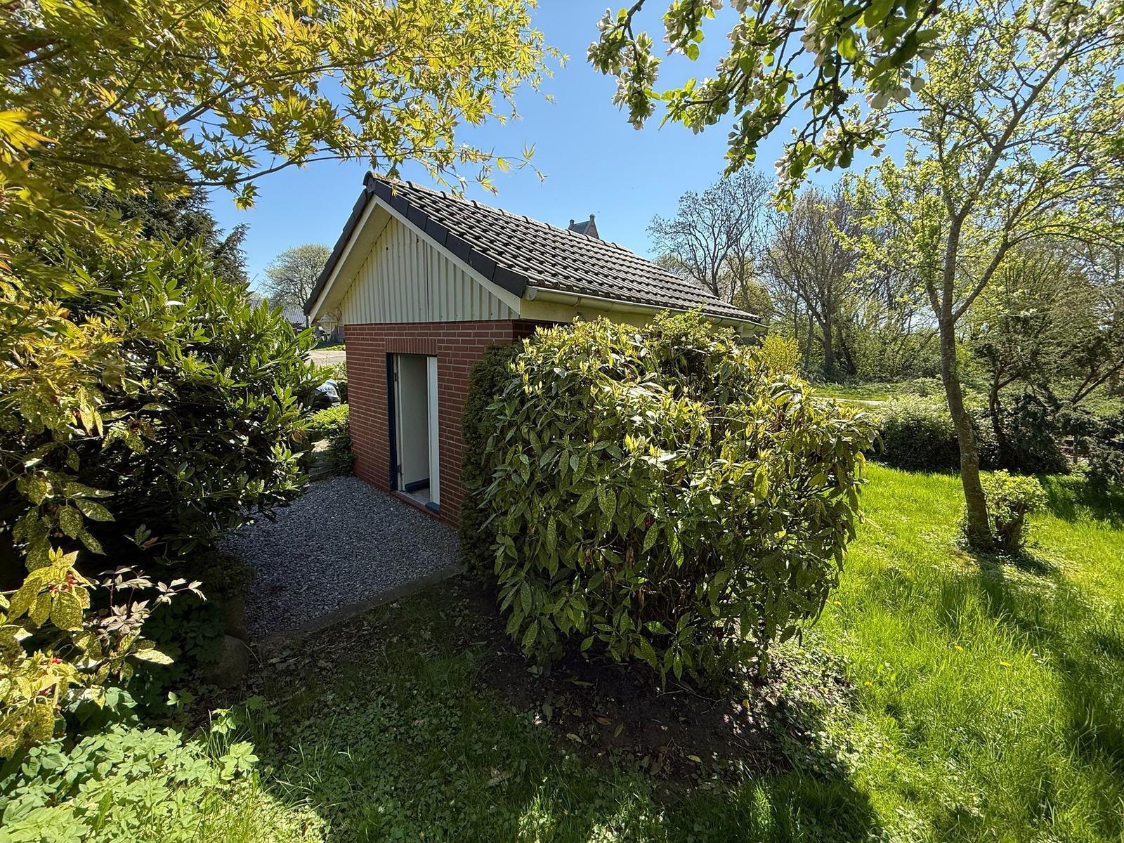 Kleines Haus mit Ziegelfassade und Dach, umgeben von Grünflächen und Bäumen.