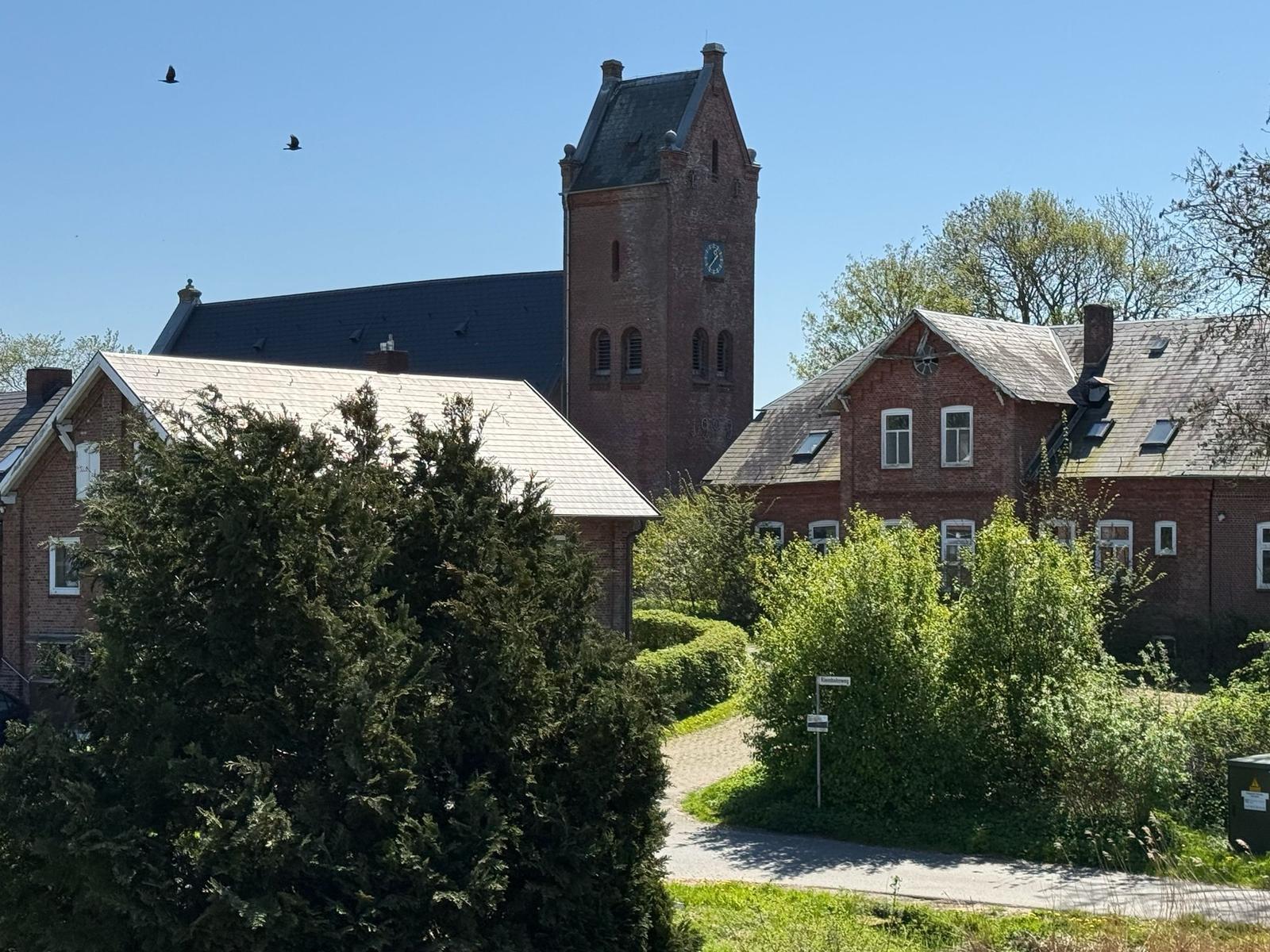 Rotes Backsteingebäude mit Turm und Dach, umgeben von Bäumen und einem Weg.