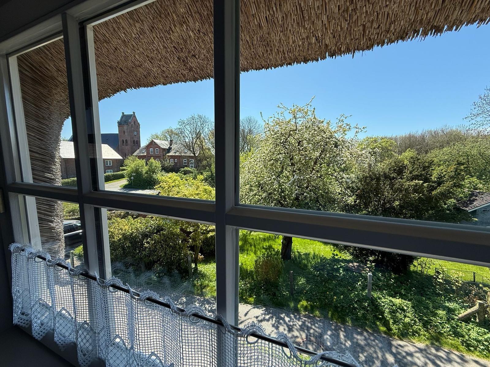 Durch das Fenster blickt man auf einen Garten mit Bäumen und ein rotes Haus mit Turm.