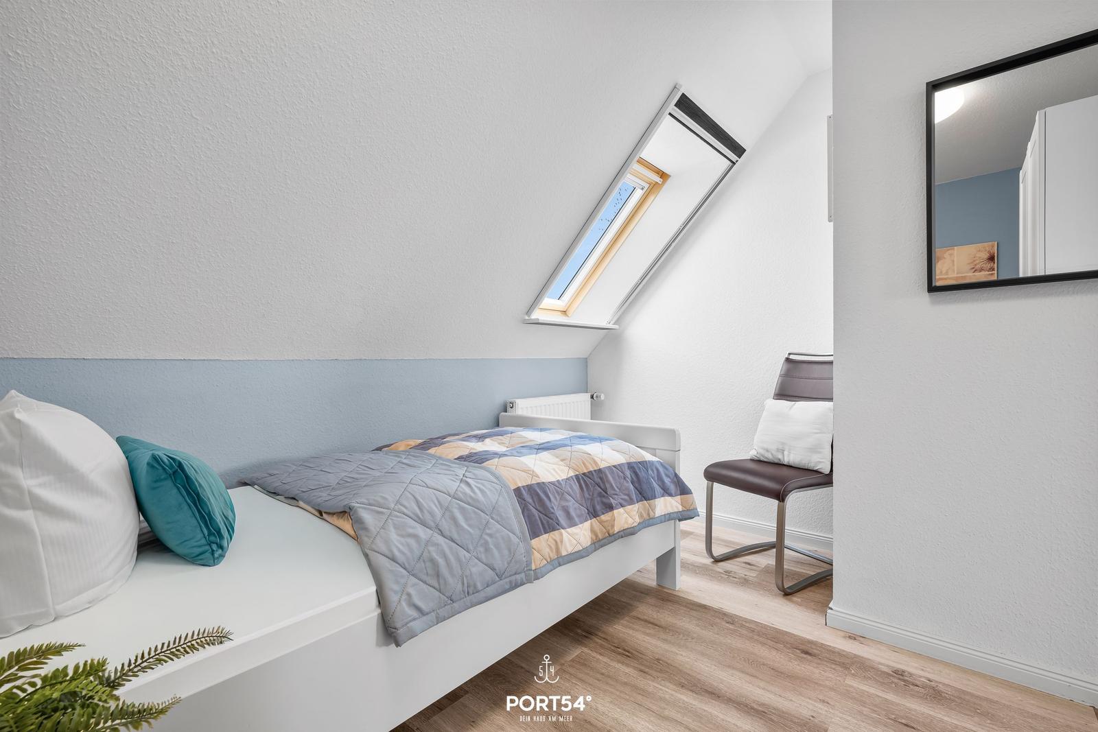 Schlafzimmer mit Dachfenster, Bett, Stuhl und Spiegel an der Wand.