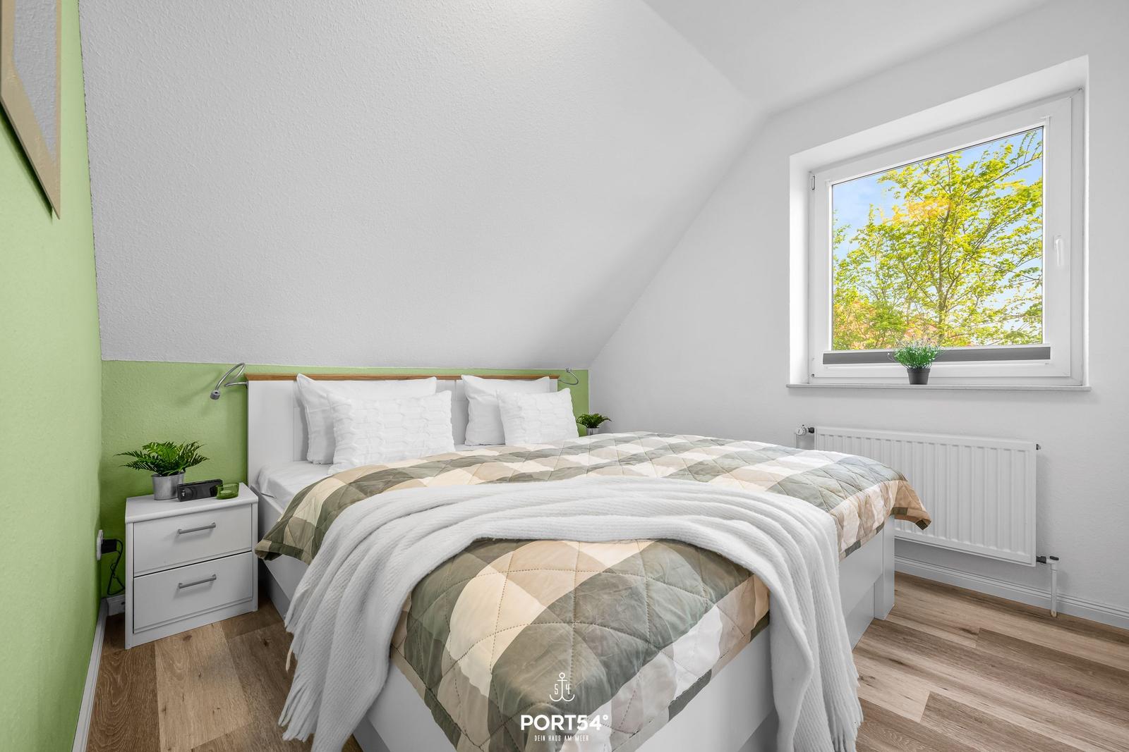 Schlafzimmer mit Dachschräge, Bett, Nachttisch und Fenster mit Blick auf Bäume.