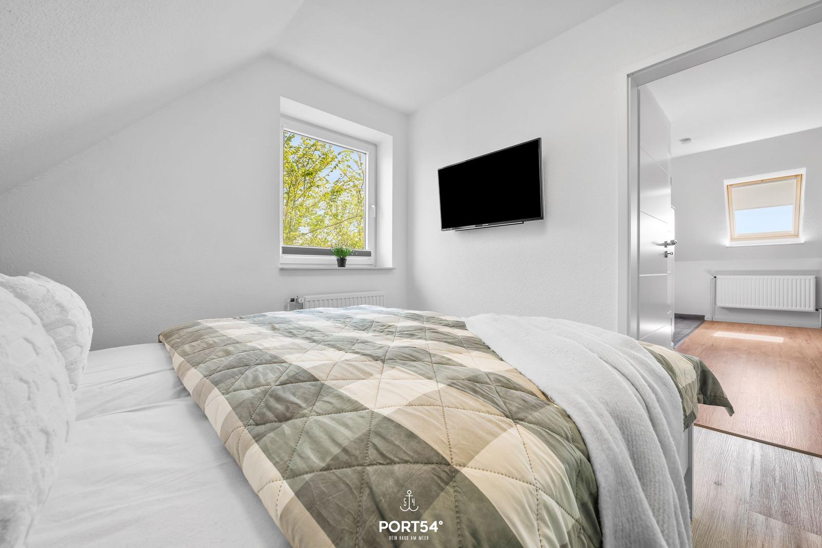 Schlafzimmer mit Bett, Fenster und Fernseher. Blick in angrenzenden Raum.