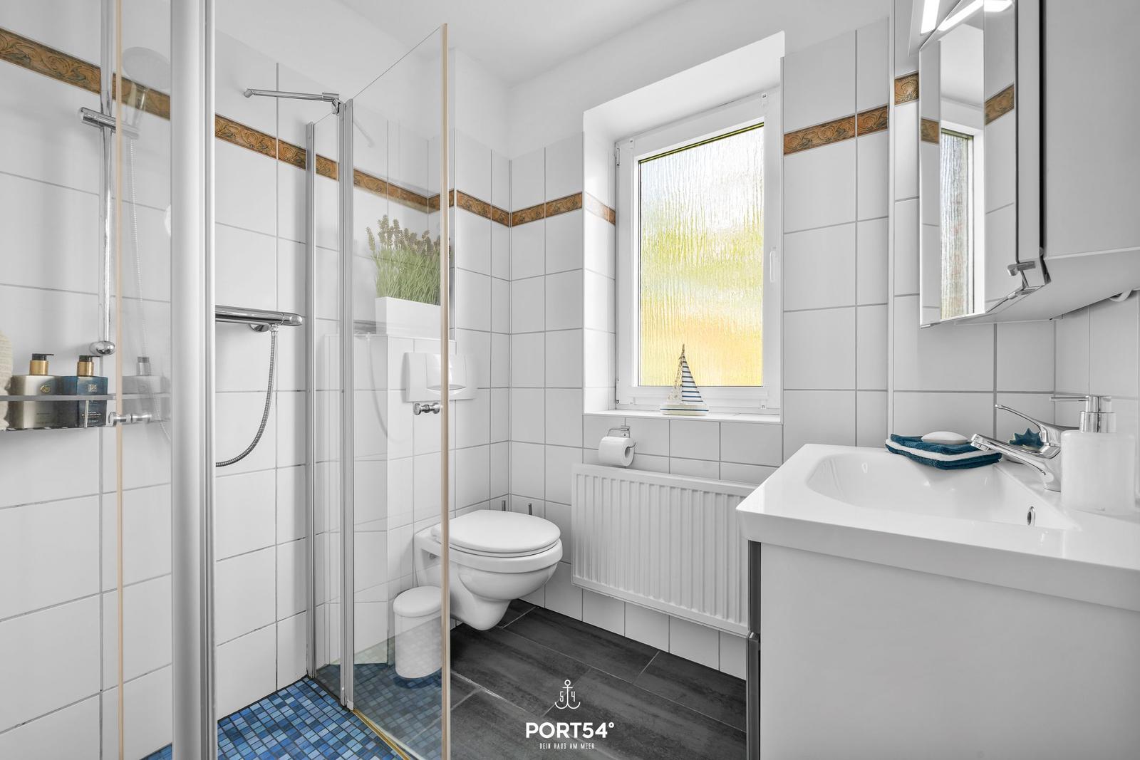 Badezimmer mit Dusche, Toilette und Waschbecken. Fenster mit getöntem Glas.