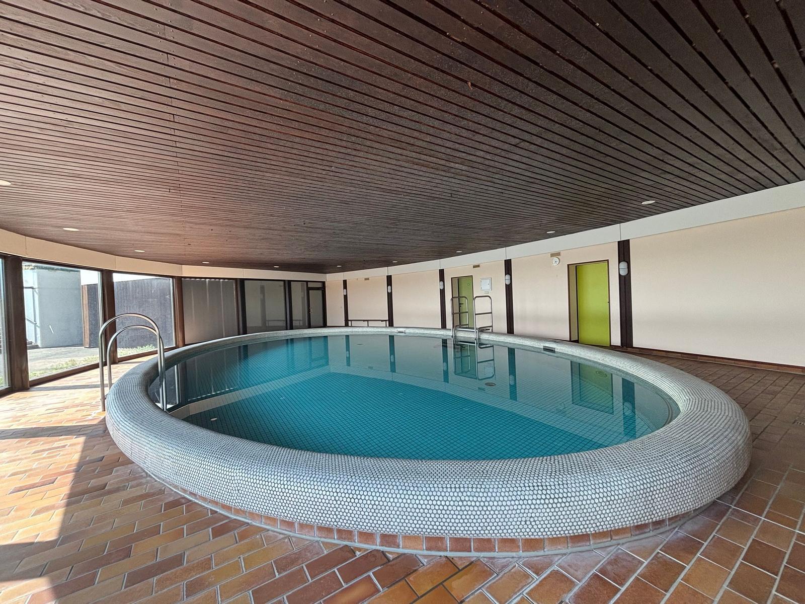 Runder Innenpool mit Mosaikrand und Holzdecke. Zwei Metalltreppen und Fenster zur Außenseite.