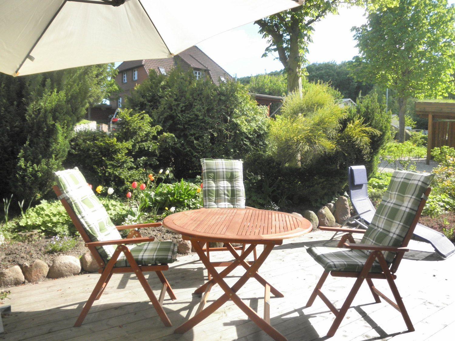 Terrasse mit Tisch, Stühlen und Sonnenschirm. Hintergrund: Garten mit Sträuchern und Haus.