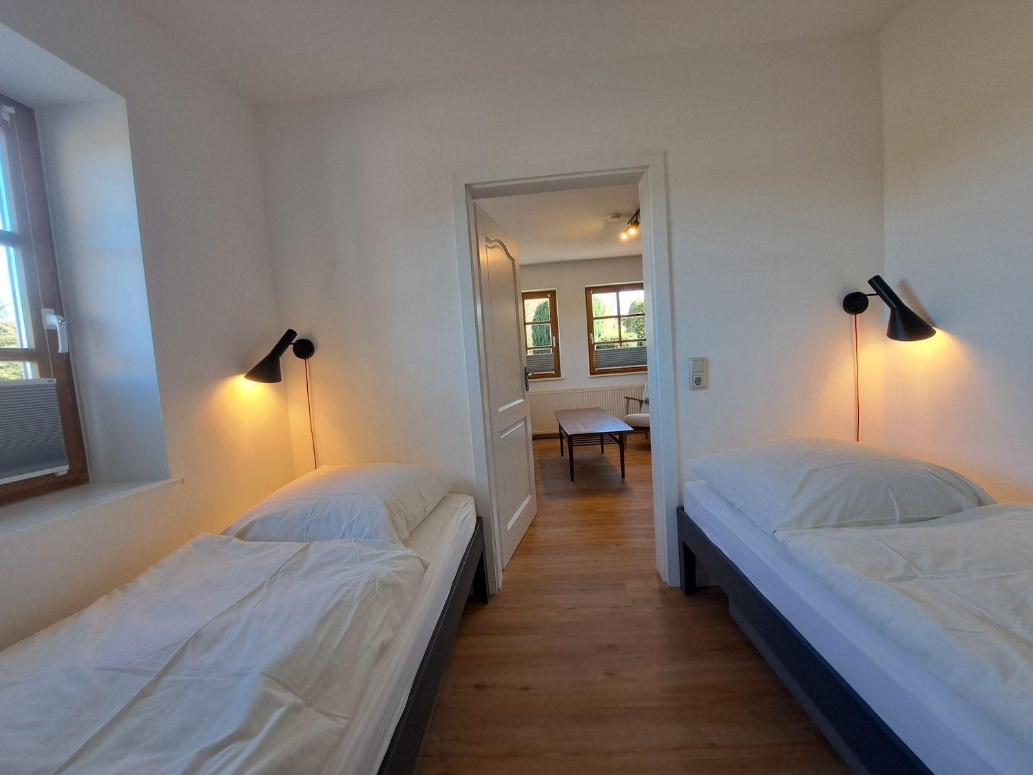 Zwei Betten mit weißen Bettwäsche, Wandlampen und Holzboden. Durchgang zu Wohnzimmer mit Tisch.