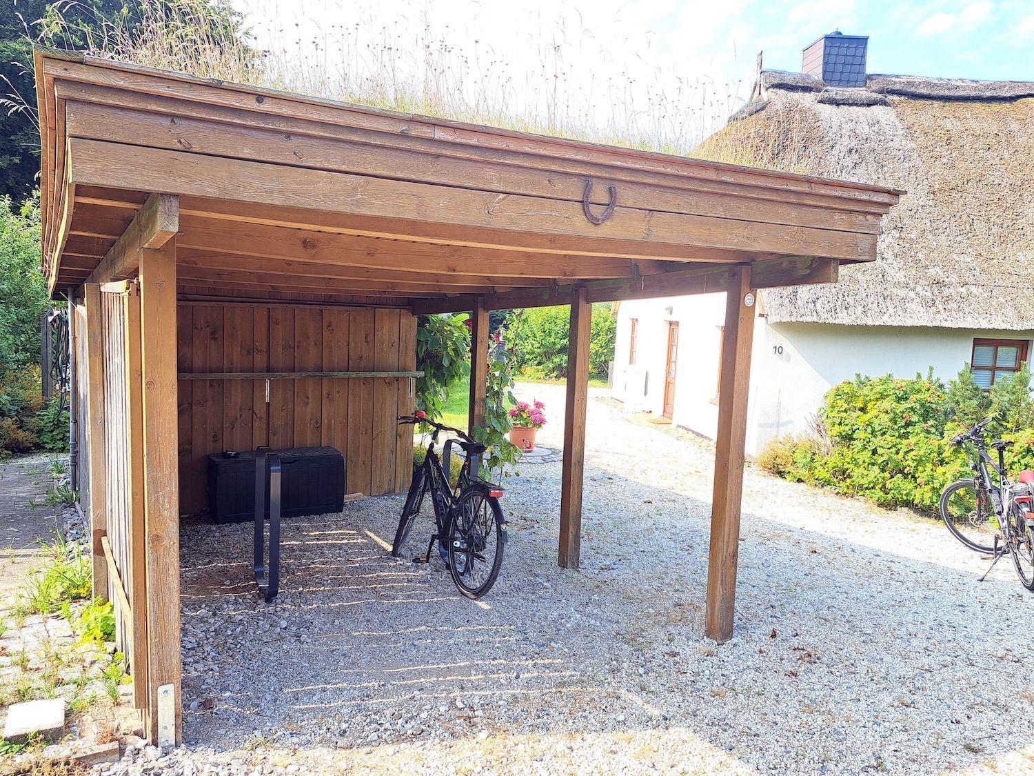 Holzüberdachung mit Fahrrädern und Schuppen neben einem Strohdachhaus.
