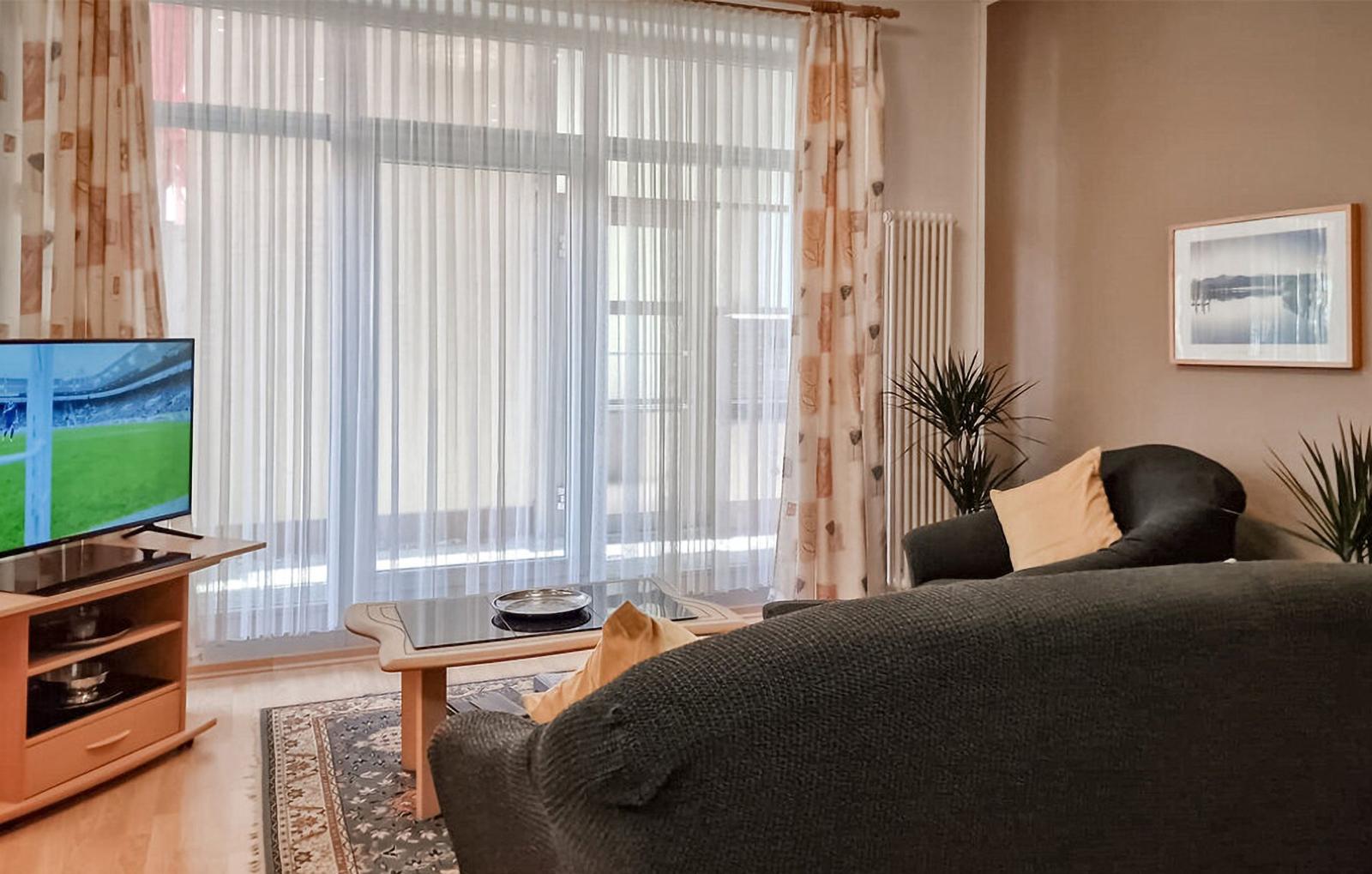Wohnzimmer mit Sofa, Fernseher und großen Fenstern mit Vorhängen.