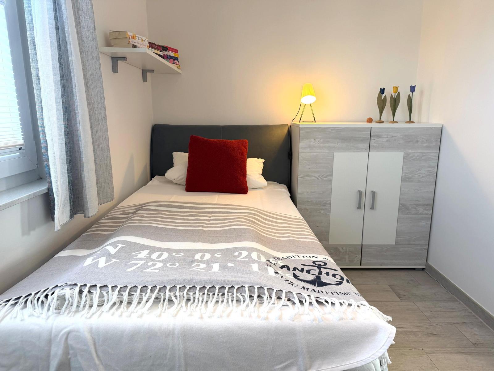 Schlafzimmer mit Bett, Nachttisch und Kleiderschrank. Auf dem Bett liegt eine Decke mit Koordinaten.