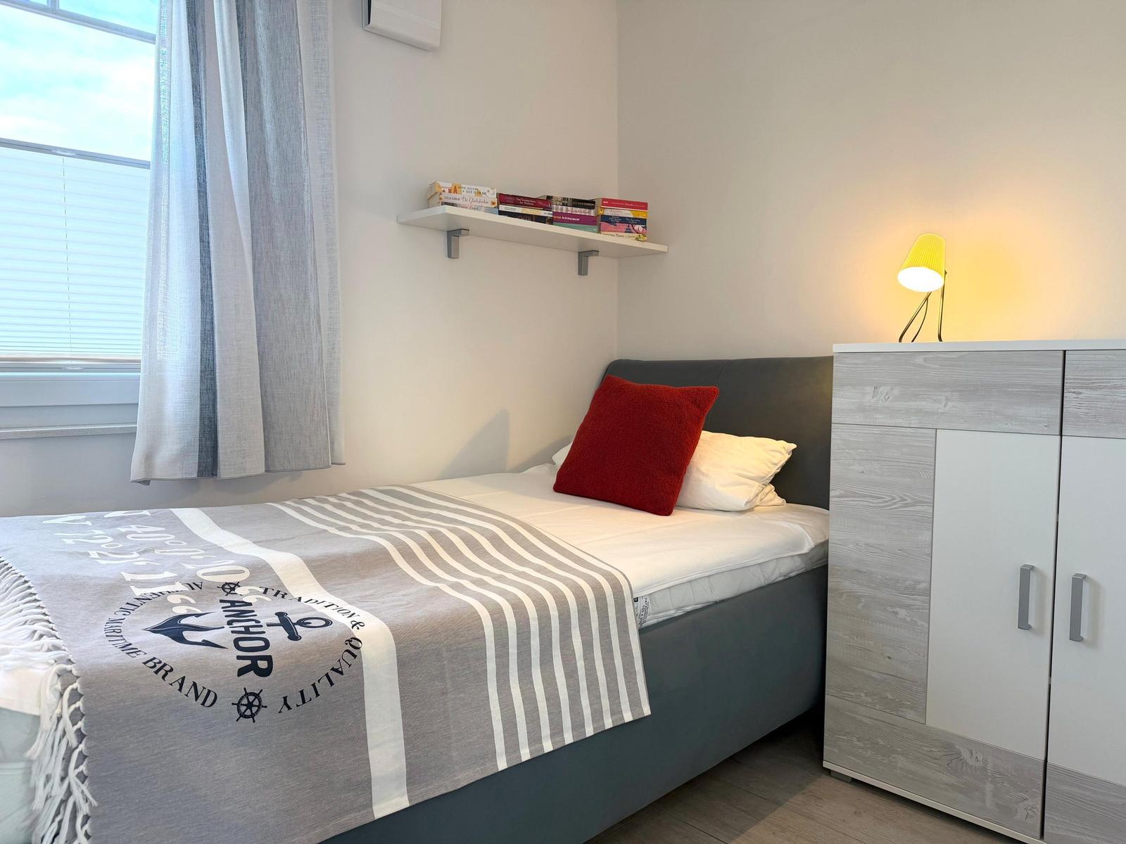 Ein Schlafzimmer mit Bett, Nachttisch und Fenster. Auf dem Bett liegt eine graue Decke mit Streifen.