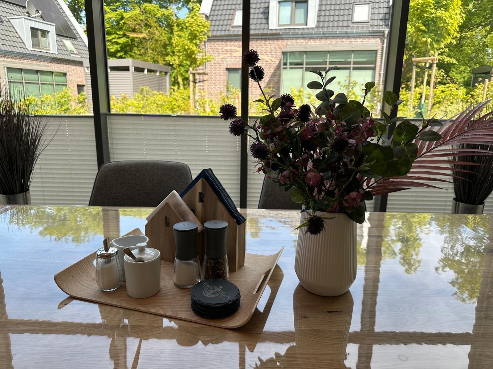 Esstisch mit Blumenvase, Gewürzdosen und Servierplatte vor großem Fenster