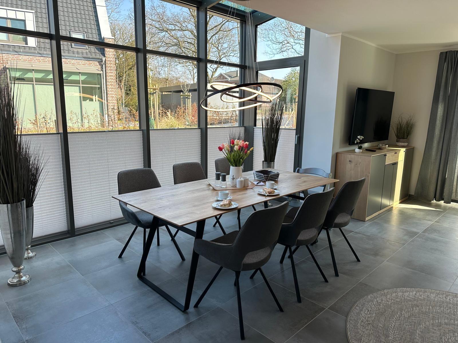 Esszimmer mit Tisch, Stühlen und großen Fenstern. Fernseher und Pflanzen sind sichtbar.