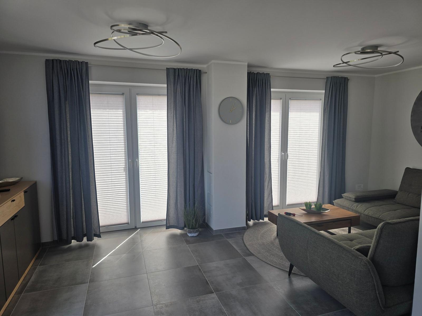Wohnzimmer mit grauen Möbeln, Fenstern und Teppich. Deckenleuchten und Wanduhr sichtbar.