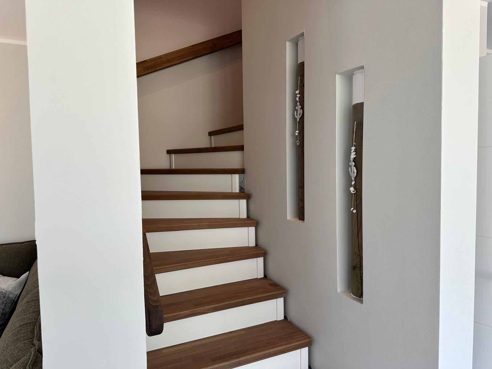 Treppe mit Holzstufen und weißen Wänden. An der Wand hängen zwei dekorative Elemente.