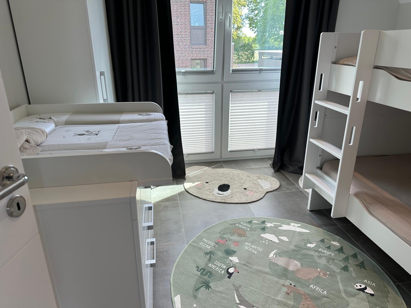Zimmer mit Kinderbett, Doppelbett und zwei Teppichen. Fenster mit Jalousien und Vorhängen.